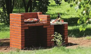 Prosty murowany grill z cegły, gotowy do przyjęcia gości. Na ruszcie już leżą smakołyki, a obok butelka wina i warzywa.