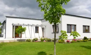 Nowoczesny bungalow z białymi ścianami i dużymi oknami, otoczony zielonym trawnikiem i kwitnącymi krzewami.