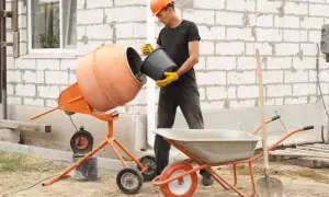 Budowlaniec w kasku wsypuje beton do betoniarki. Obok stoi taczka i łopata.