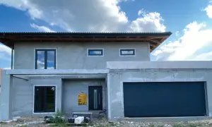 Budowa domu z szarym tynkiem, czarnymi oknami i bramą garażową pod błękitnym niebem.