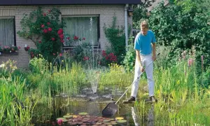 Mężczyzna pielęgnuje bujne oczko wodne w ogrodzie, usuwając liście siatką. Wokół kwitną róże i inne kwiaty.