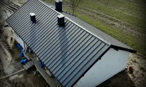 Nowy, ciemnoszary dach z blachy na domu jednorodzinnym. Widać kominy i otoczenie budowy. Dobry koszt dachu to podstawa.