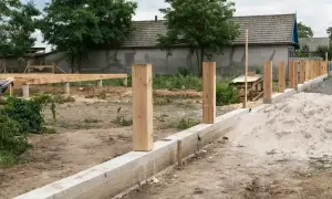 Solidny fundament pod ogrodzenie z betonowym pasem i drewnianymi słupkami, w tle budowa domu.