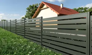 Nowoczesny montaż ogrodzenia panelowego cena. Elegancki płot z poziomych desek, otaczający dom z czerwonym dachem, na tle zielonej trawy i błękitnego nieba.