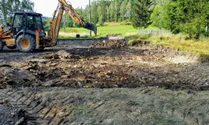 Koparka pracuje, tworząc wykopy pod fundamenty. Teren jest błotnisty, z gruzem i kamieniami.