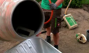 Osoba w rękawiczkach miesza beton w betoniarce, obok stoi taczka i worek cementu.