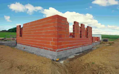 Budowa domu w toku: czerwone ściany z pustaków piętrzą się na betonowym fundamencie. W tle zielone wzgórza i błękitne niebo z chmurami. Czas budowy domu zależy od wielu czynników.