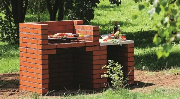 Prosty murowany grill z cegły, gotowy do przyjęcia gości. Na ruszcie już leżą smakołyki, a obok butelka wina i warzywa.