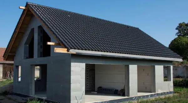 Dom w stanie surowym otwartym, z betonowych pustaków, z dachem pokrytym czarną dachówką. Widać otwory okienne i drzwiowe.