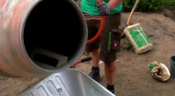 Osoba w rękawiczkach miesza beton w betoniarce, obok stoi taczka i worek cementu.