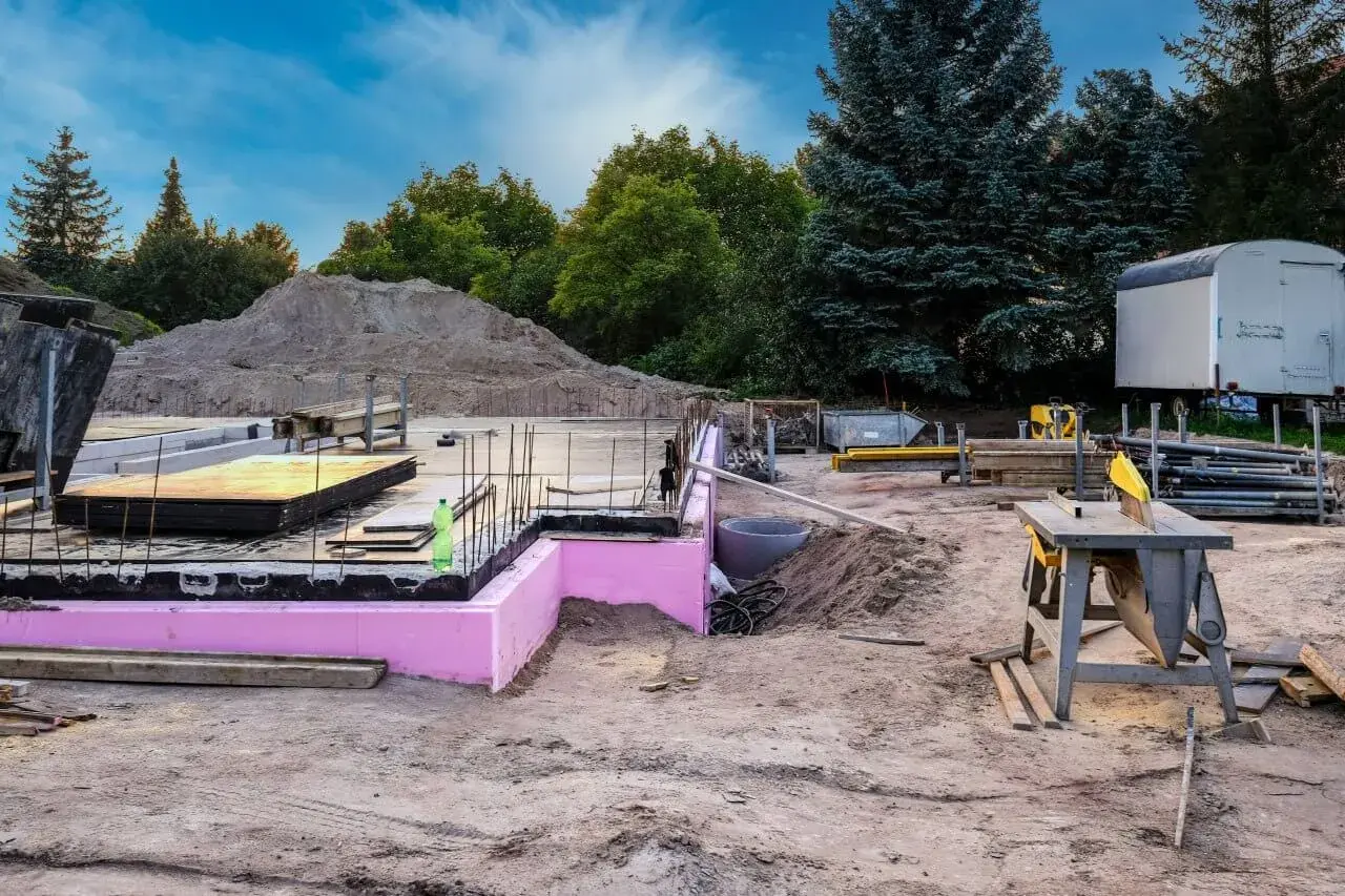 Budowa w stanie zerowym: fundamenty z różową izolacją, zbrojenie, sterta piasku i sprzęt budowlany.