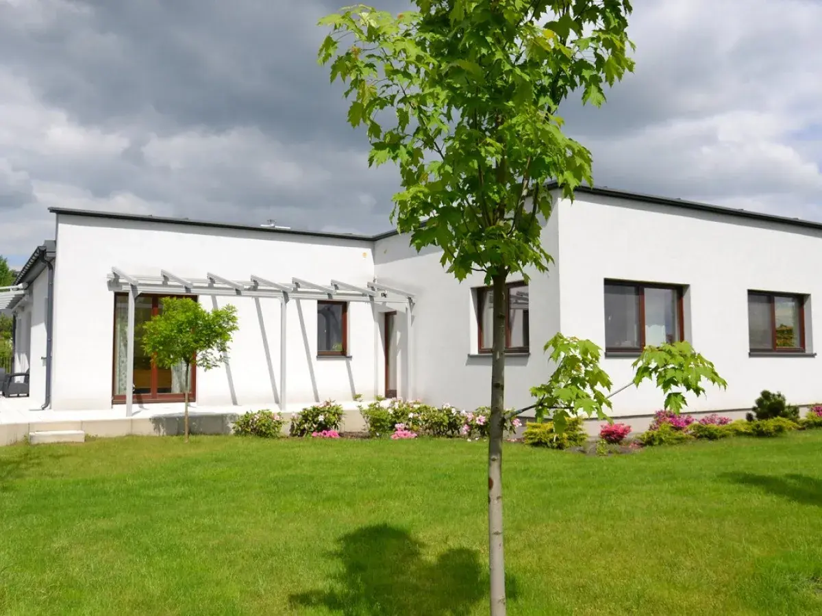 Nowoczesny bungalow z białymi ścianami i dużymi oknami, otoczony zielonym trawnikiem i kwitnącymi krzewami.