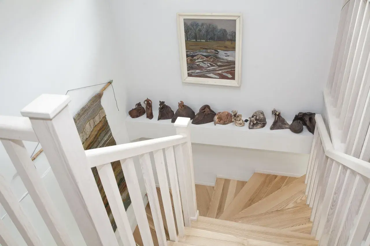 Jasna klatka schodowa z drewnianymi stopniami i balustradą, na półce nad nią rzeźby i obraz.