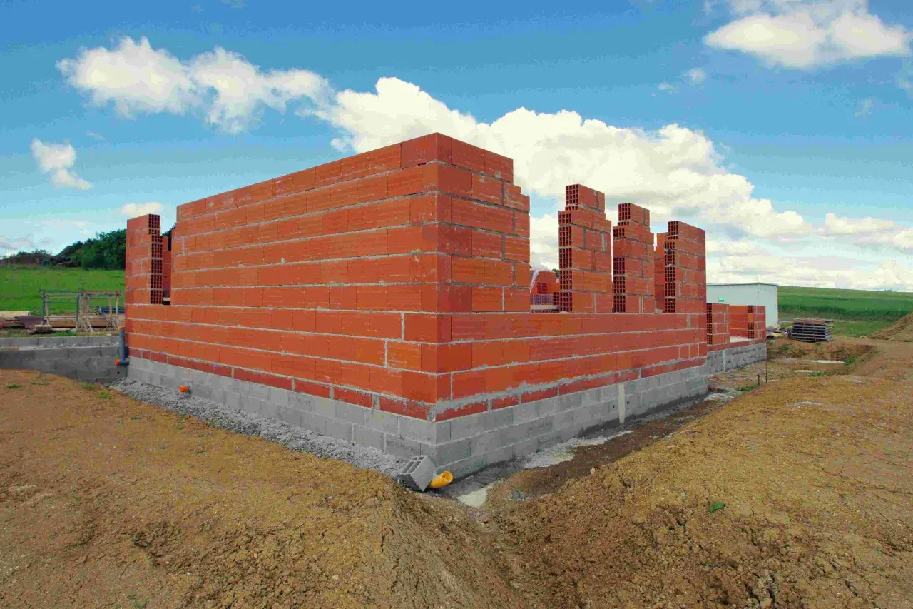 Budowa domu w toku: czerwone ściany z pustaków piętrzą się na betonowym fundamencie. W tle zielone wzgórza i błękitne niebo z chmurami. Czas budowy domu zależy od wielu czynników.