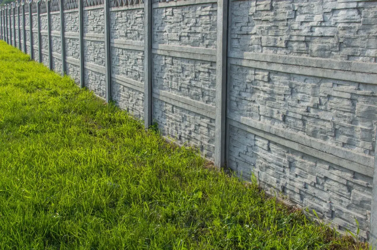 Nowy, betonowy płot z fakturą kamienia, otoczony soczystą zieloną trawą. Idealny do malowania i zabezpieczenia.