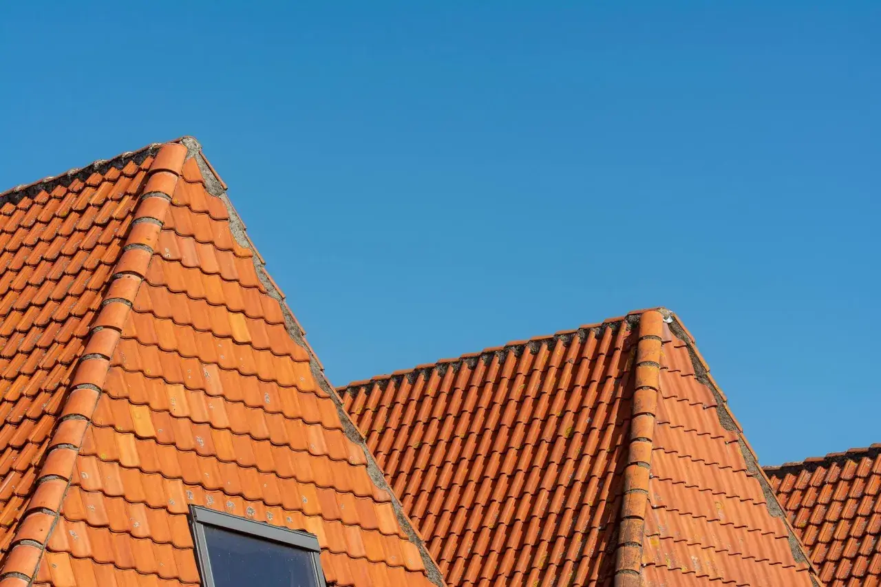Pomarańczowy dach wielospadowy z dachówki ceramicznej pod błękitnym niebem. Widoczny fragment okna dachowego.