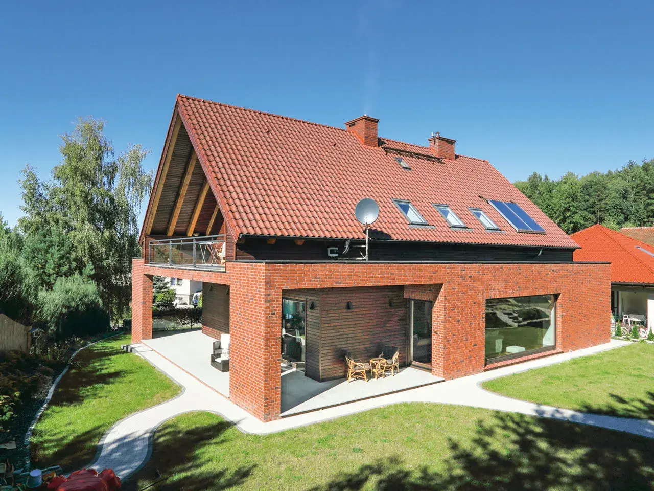 Nowoczesny dom z dachem dwuspadowym 100m2, ceglaną elewacją i dużymi oknami. W ogrodzie ścieżki i zieleń.