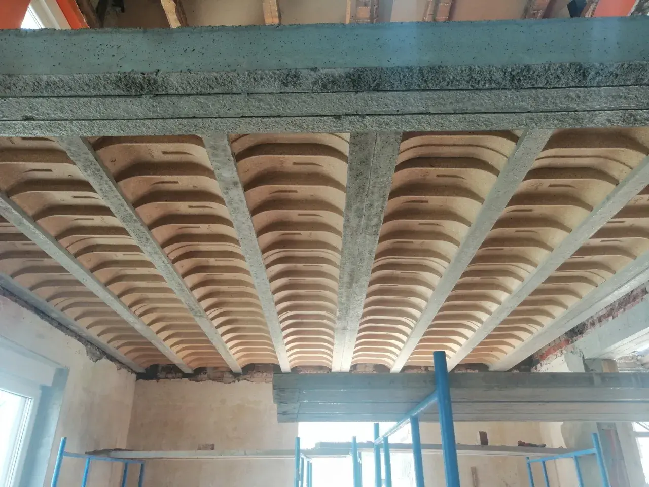 Widok od dołu na budowę, gdzie widoczne są różne rodzaje stropów: betonowe belki i płyty, a także pustaki ceramiczne tworzące łuki.