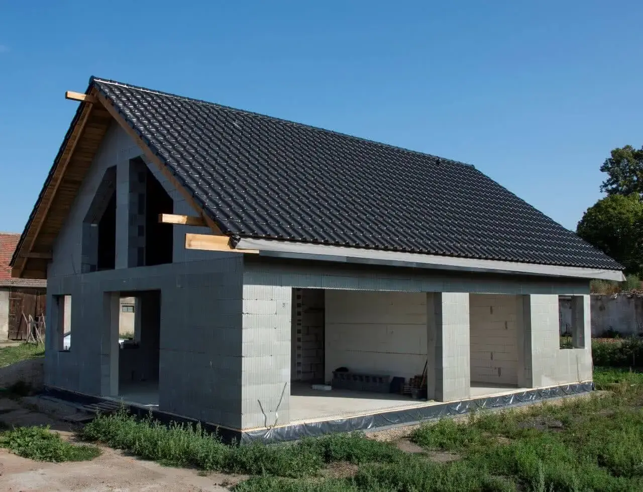 Dom w stanie surowym otwartym, z betonowych pustaków, z dachem pokrytym czarną dachówką. Widać otwory okienne i drzwiowe.