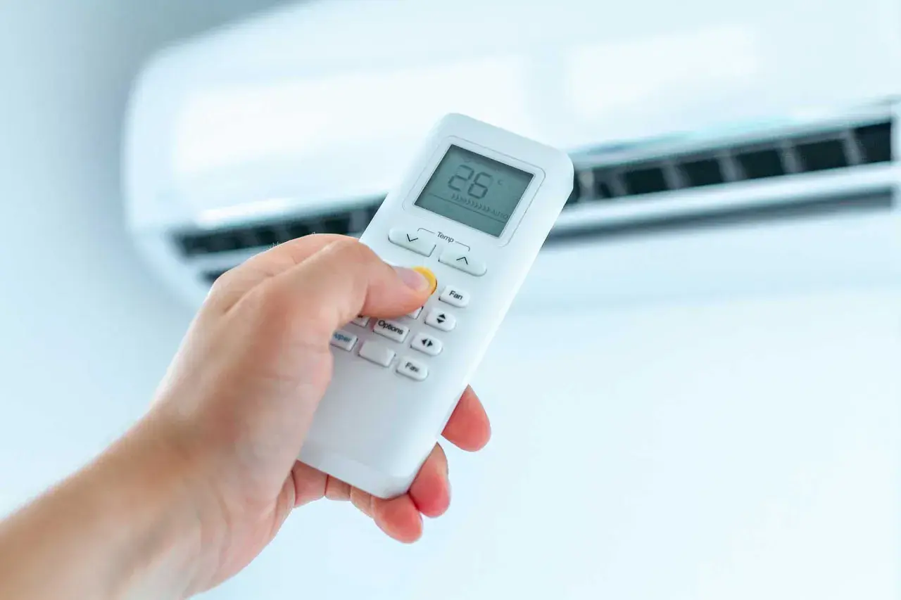 Dłoń trzymająca pilot do klimatyzacji, ustawiająca ogrzewanie. Na ekranie widoczna temperatura 26°C.