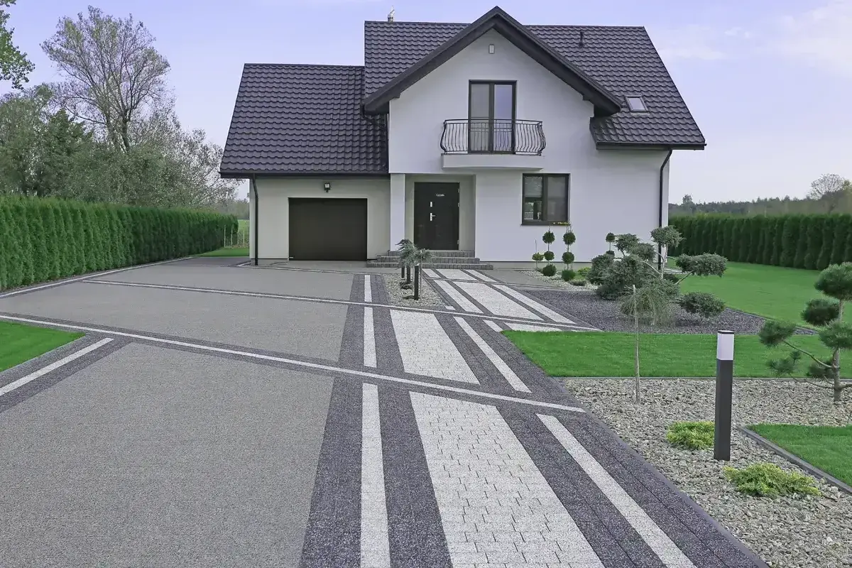 Nowoczesny podjazd do domu z geometrycznymi wzorami z kostki brukowej, prowadzący do białego domu z garażem i zadbanym ogrodem.