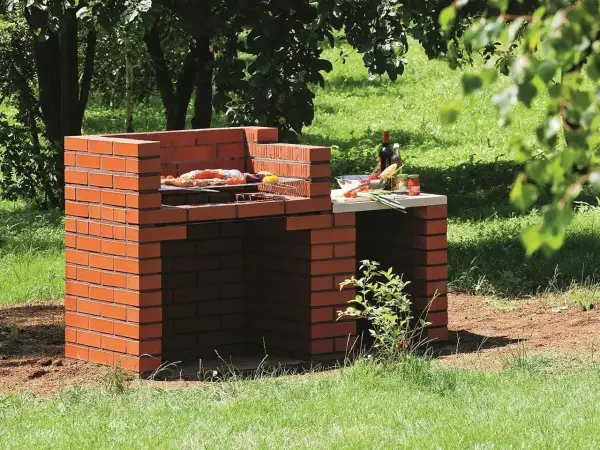 Prosty murowany grill z cegły, gotowy do przyjęcia gości. Na ruszcie już leżą smakołyki, a obok butelka wina i warzywa.