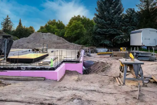 Budowa w stanie zerowym: fundamenty z różową izolacją, zbrojenie, sterta piasku i sprzęt budowlany.