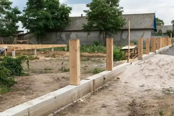 Solidny fundament pod ogrodzenie z betonowym pasem i drewnianymi słupkami, w tle budowa domu.