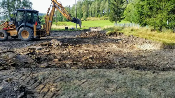 Koparka pracuje, tworząc wykopy pod fundamenty. Teren jest błotnisty, z gruzem i kamieniami.